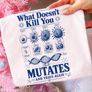 Puede incluir: Una camiseta blanca con un estampado gráfico azul que incluye el texto "What Doesn't Kill You MUTATES AND TRIES AGAIN". El diseño muestra un diagrama de mutación celular de cuatro etapas, una doble hélice de ADN y estructuras químicas.