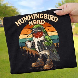 Könnte beinhalten: Schwarzes T-Shirt mit einem Cartoon-Kolibri, der eine Brille trägt und ein Fernglas und eine Kaffeetasse hält. Der Text "HUMMINGBIRD NERD" ist über dem Vogel gewölbt, mit einem Sonnenuntergangsdesign im Hintergrund.