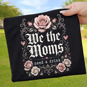 Op de afbeelding: Zwart t-shirt met de tekst "We the Moms need a drink" in een gotisch lettertype, omringd door roze rozen en decoratieve krullen. Twee roze harten flankeren de tekst.