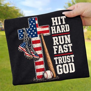 Puede incluir: Una camiseta negra con un gráfico de una cruz con el patrón de la bandera estadounidense, un par de zapatillas de béisbol, un bate de madera y una pelota. El texto HIT HARD, RUN FAST, TRUST GOD aparece en letras blancas desgastadas.