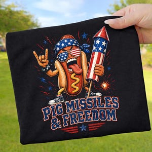 Könnte beinhalten: Ein schwarzes T-Shirt mit einer Grafik eines Hot-Dog-Charakters, der ein Bandana und eine Sonnenbrille mit US-Flaggenmuster trägt. Die Figur hält einen Feuerwerkskörper und macht eine Rock-Geste. Text: PIG MISSILES & FREEDOM.