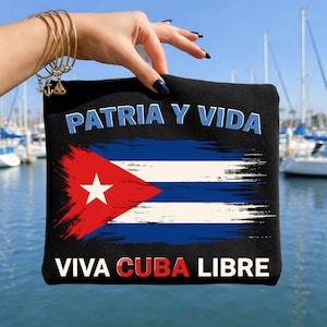 Puede incluir: Bolsa negra con el diseño de la bandera cubana, que incluye un triángulo rojo con una estrella blanca, rayas azules y blancas, y las frases "PATRIA Y VIDA" y "VIVA CUBA LIBRE". El fondo es un puerto deportivo.