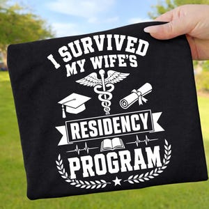 Könnte beinhalten: Ein schwarzes T-Shirt mit weißem Aufdruck, der I SURVIVED MY WIFE'S RESIDENCY PROGRAM lautet. Das Design enthält ein medizinisches Caduceus-Symbol, eine Doktorhut, ein zusammengerolltes Diplom, ein offenes Buch und eine Herzschlag-Grafik.
