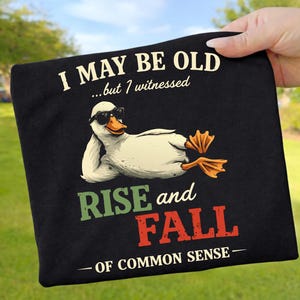 Könnte beinhalten: Schwarzes T-Shirt mit einer Cartoon-Ente mit Sonnenbrille. Der Text auf dem Shirt lautet: "I MAY BE OLD...but I witnessed RISE and FALL - OF COMMON SENSE -" in Weiß, Grün und Rot.