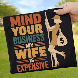 Pode incluir: Camiseta preta com o texto "MIND YOUR BUSINESS MY WIFE IS EXPENSIVE" em laranja, amarelo e turquesa. Uma mulher de desenho animado em um vestido e salto alto carrega uma bolsa de dinheiro.