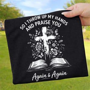 Op de afbeelding: Zwart t-shirt met een witte afbeelding van een kruis, rozen, vlinders en een open boek. De tekst luidt "So I throw up my hands and praise you" en "Again & Again".