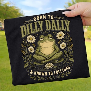 May include: A black t-shirt featuring a graphic of a green frog sitting in a lotus position on a lily pad. The frog is surrounded by white daisies and green foliage. The text reads BORN TO DILLY DALLY & KNOWN TO LOLLYGAG in a light green font.
