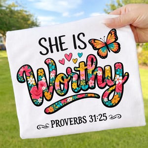 Könnte beinhalten: Weißes T-Shirt mit den Worten "SHE IS WORTHY" in einem floralen Design. Ein bunter Schmetterling und Herzen befinden sich über dem Wort "Worthy". Der Text "PROVERBS 31:25" steht darunter.