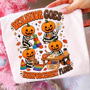 May include: A white t-shirt featuring a graphic of four skeletons with pumpkin heads playing with toys. The text reads "BEHAVIOR GOES" at the top and "REINFORCEMENT FLOWS" at the bottom. One skeleton holds a "GOOD JOB!" card, and a box is labeled "CALM FOCUS FUN".