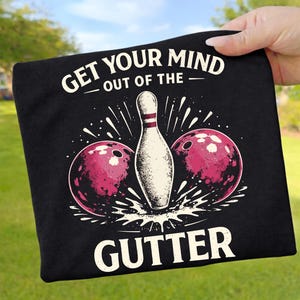 Könnte beinhalten: Schwarzes T-Shirt mit Bowling-Motiv. Das Design zeigt eine weiße Bowlingkegel, zwei weinrote Bowlingkugeln und den Text "GET YOUR MIND OUT OF THE GUTTER" in Weiß.