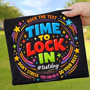 Könnte beinhalten: Schwarzes T-Shirt mit einem farbenfrohen Grafikdesign. Das Design enthält den Text "ROCK THE TEST TIME TO LOCK IN" in verschiedenen Farben, umgeben von Sternen und Regenbogenbögen. Der Text "DON'T STRESS YOU CAN DO IT DO YOUR BEST" ist ebenfalls enthalten.