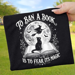 May include: A black t-shirt featuring a white graphic of an open book with a black cat sitting on the pages. Above the book is a full moon with a witch flying on a broomstick and bats. The text reads: TO BAN A BOOK IS TO FEAR ITS MAGIC.
