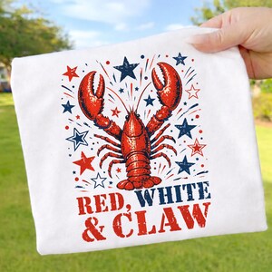 Könnte beinhalten: Weißes T-Shirt mit einer roten Hummer-Illustration, blauen und roten Sternen und Feuerwerk. Der Text "RED, WHITE & CLAW" ist darunter in Rot und Blau gedruckt.