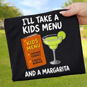 May include: A black t-shirt featuring white text that reads I'LL TAKE A KIDS MENU AND A MARGARITA. The graphic includes an orange menu labeled KIDS MENU with CHICKEN TENDIES and CHEESE BURGER listed, alongside a green margarita with a lime slice.