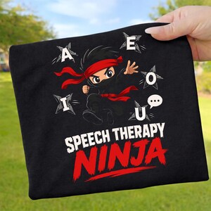 Puede incluir: Camiseta negra con un diseño de un ninja de dibujos animados. El ninja lleva una cinta para la cabeza y una faja rojas. El texto "SPEECH THERAPY NINJA" está en blanco y rojo. Las letras A, E, I, O y U están en el fondo.