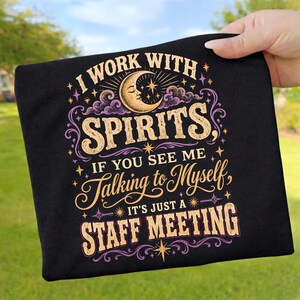 Könnte beinhalten: Ein schwarzes T-Shirt mit einem Grafikdesign aus einer Mondsichel, Sternen und violetten Wolken. Der Text lautet: I WORK WITH SPIRITS, IF YOU SEE ME TALKING TO MYSELF, IT'S JUST A STAFF MEETING. Die Schrift besteht aus einer Mischung aus fetten und Schreibschrift-Stilen.