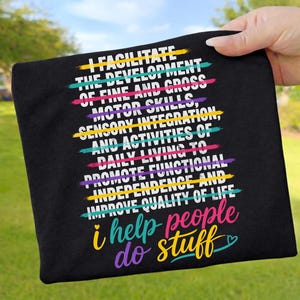 Könnte beinhalten: Schwarzes T-Shirt mit dem Text "I FACILITATE THE DEVELOPMENT... i help people do stuff" in Weiß, mit bunten Linien durch den Text. Das T-Shirt wird von einer Person gehalten.