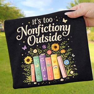 Könnte beinhalten: Schwarzes T-Shirt mit dem Text "it's too Nonfictiony Outside" in weißer Schrift. Das Design zeigt bunte Bücher, Sonnenblumen und Schmetterlinge. Eine Hand hält das Shirt vor einem grünen Hintergrund.