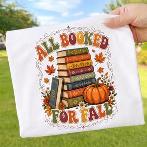 Könnte beinhalten: Ein weißes T-Shirt mit einer Grafik aus einem Stapel bunter Bücher und einem orangefarbenen Kürbis. Der Text "ALL BOOKED FOR FALL" ist in einer Retro-Schrift über und unter dem Stapel gedruckt, umgeben von herbstlichen Blättern und Blumen.