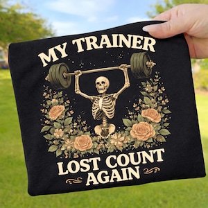 May include: Black t-shirt featuring the text "MY TRAINER" above a skeleton lifting weights, with floral details and the text "LOST COUNT AGAIN". The design includes a barbell with weights and peach-coloured roses.