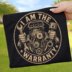 Könnte beinhalten: Ein schwarzes T-Shirt mit einer beigefarbenen kreisförmigen Grafik eines Automotors, wobei eine Hand einen Schraubenschlüssel und die andere einen Kolben hält. Der Text "I AM THE WARRANTY" ist in einer abgenutzten Schriftart um das Design gedruckt.