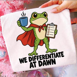 Op de afbeelding: Een wit t-shirt met een illustratie van een groene kikker in een rode cape, die een blauwe koffiemok en een klembord met een checklist vasthoudt. Onder de kikker staat de tekst WE DIFFERENTIATE AT DAWN in dikke, zwarte hoofdletters gedrukt.