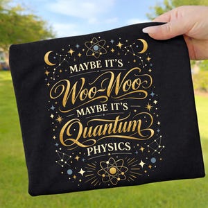 May include: Black t-shirt with gold lettering and celestial graphics. The text reads "Maybe it's Woo-Woo, Maybe it's Quantum Physics." The design includes stars, constellations, and atom symbols. The shirt is held up by a hand.