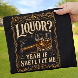 Könnte beinhalten: Schwarzes T-Shirt mit dem Text "LIQUOR? YEAH IF SHE'LL LET ME" in Gold und Weiß. Das Design zeigt ein Whiskeyglas mit Eis, eine brennende Zigarre und dekorative Verzierungen.