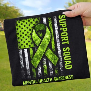 Könnte beinhalten: Schwarzes Quadrat mit einem verwitterten Design der amerikanischen Flagge in Grün und Weiß. Ein grünes Band ist über der Flagge zentriert, mit den Worten "SUPPORT SQUAD" und "MENTAL HEALTH AWARENESS" in Grün.
