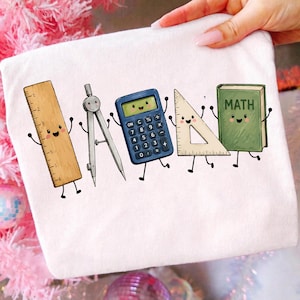 Può includere: Una t-shirt bianca con una stampa grafica di cinque articoli scolastici antropomorfi: un righello beige, un compasso argentato, una calcolatrice blu, una squadra trasparente e un libro verde con la parola MATH sulla copertina.