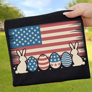 Könnte beinhalten: Schwarzes T-Shirt mit einem Design der amerikanischen Flagge im Used-Look. Das Design umfasst weiße Kaninchensilhouetten, dekorierte Ostereier in Rot, Weiß und Blau sowie Sterne. Das T-Shirt wird von einer Hand gehalten.