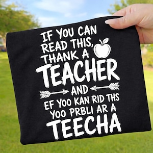 Könnte beinhalten: Schwarzes T-Shirt mit weißem Text: „IF YOU CAN READ THIS, THANK A TEACHER“ mit einer Apfelgrafik. Auf der Unterseite des T-Shirts steht „EF YOO KAN RID THS YOO PRBLI AR A TEECHA“.