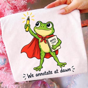 May include: A white t-shirt featuring a green frog wearing a red cape, holding a yellow highlighter and a spiral-bound notebook labeled NOTES. Below the frog, the text reads We annotate at dawn with small decorative lines on each side.