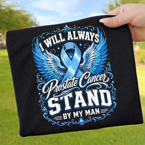Könnte beinhalten: Schwarzes T-Shirt mit blauem Band und Engelsflügel-Design. Der Text lautet "I Will Always Prostate Cancer Stand By My Man". Das Design ist in Weiß und Blau gehalten.