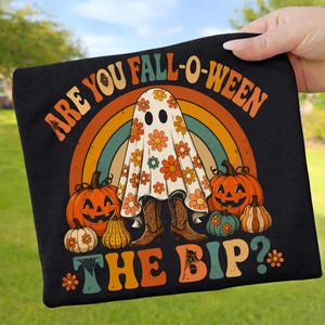 Könnte beinhalten: Ein schwarzes T-Shirt mit einer Grafik eines Geistes, der ein Laken mit Blumenmuster und braune Cowboystiefel trägt. Das Design umfasst Kürbisse, einen Regenbogen und den Text ARE YOU FALL-O-WEEN THE BIP? in Retro-Farben wie Orange, Petrol und Gelb.