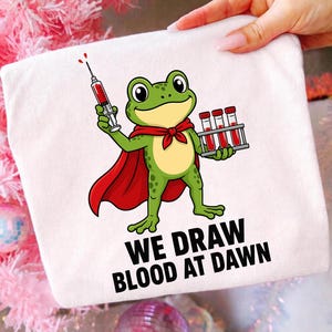 Könnte beinhalten: Ein weißes T-Shirt mit einem grünen Frosch in einem roten Umhang, der eine Spritze in der einen und einen Ständer mit drei mit roter Flüssigkeit gefüllten Reagenzgläsern in der anderen Hand hält. Darunter steht WE DRAW BLOOD AT DAWN in Schwarz.