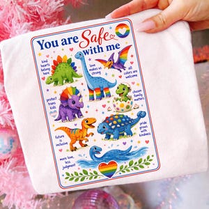 Könnte beinhalten: Ein weißes T-Shirt mit einer bunten Grafik verschiedener Dinosaurier und dem Text "You are Safe with me". Das Design enthält Sätze wie "kind hearts belong here", "protect trans kids" und "more love, less judgment" sowie regenbogenfarbene Herzen.