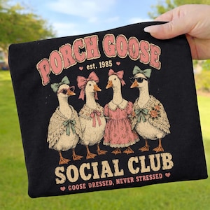 Könnte beinhalten: Schwarzes T-Shirt mit dem Text "PORCH GOOSE SOCIAL CLUB est. 1985". Das Design zeigt vier Gänse mit Accessoires wie Sonnenbrillen und Schleifen. Der Satz "GOOSE DRESSED, NEVER STRESSED" ist ebenfalls aufgedruckt.