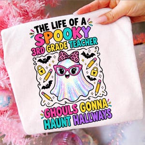 Könnte beinhalten: Ein weißes T-Shirt mit einer bunten Grafik und dem Text THE LIFE OF A SPOOKY 3RD GRADE TEACHER sowie GHOULS GONNA HAUNT HALLWAYS. Das Motiv zeigt einen holografischen Geist mit rosa Brille und Leopardenmuster-Schleife, Bleistiften und Fledermäusen.