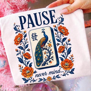 May include: A white t-shirt featuring a graphic print with the word PAUSE at the top and never mind at the bottom. The center shows a mahjong tile with a peacock illustration and Chinese character, surrounded by orange flowers and blue vines.