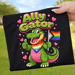 Könnte beinhalten: Schwarzes T-Shirt mit einem Cartoon-Alligator und dem Text "Ally Gator". Der grüne Alligator trägt ein Regenbogen-Bandana und hält eine Pride-Flagge. Rosa Herzen und Sterne umgeben den Alligator.