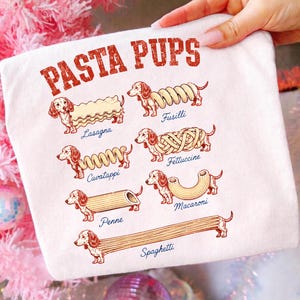 Könnte beinhalten: Ein weißes T-Shirt mit dem roten Schriftzug PASTA PUPS und Illustrationen von Dackeln, die mit verschiedenen Nudelformen kombiniert sind, beschriftet als Lasagna, Fusilli, Cavatappi, Fettuccine, Penne, Macaroni und Spaghetti in blauer Schreibschrift.