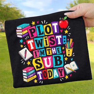 Może przedstawiać: Czarna koszulka z kolorową grafiką i napisem "PLOT TWIST I'M THE SUB TODAY". Wzór zawiera stosy książek, notatnik z długopisem, przybornik z przyborami do pisania, czerwone jabłko oraz małe żółte gwiazdki.