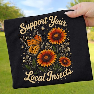 Könnte beinhalten: Schwarze Tragetasche mit einem Monarchfalter und orangefarbenen Blumen mit dem Text "Support Your Local Insects". Der Schmetterling hat orange und schwarze Flügel. Die Blumen haben orangefarbene Blütenblätter und dunkle Zentren.
