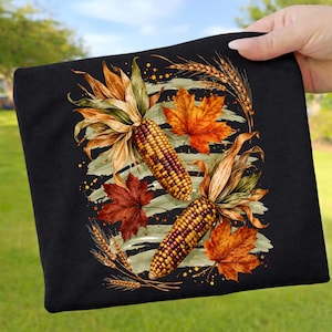 Könnte beinhalten: Ein schwarzes T-Shirt mit einem Grafikdruck von zwei mehrfarbigen Maiskolben, orangefarbenen und roten Ahornblättern sowie Weizenähren. Das Design ist auf einem hellgrünen Pinselstrich-Hintergrund mit verstreuten gelben und orangefarbenen Punkten angeordnet.