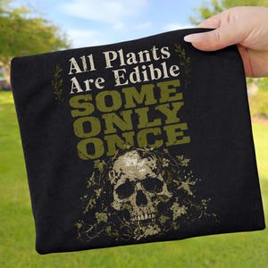 Könnte beinhalten: Schwarzes T-Shirt mit dem Text "All Plants Are Edible, Some Only Once" in Weiß und Olivgrün. Unter dem Text befindet sich eine Totenkopf-Grafik, umgeben von olivgrünen Blättern.