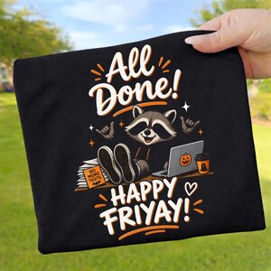 Könnte beinhalten: Ein schwarzes T-Shirt mit einer Grafik eines Waschbären, der an einem Schreibtisch mit Laptop, einem Stapel Papiere mit der Aufschrift NO MORE TESTS und einem Kaffeebecher sitzt. Der Text lautet ALL DONE! und HAPPY FRIYAY! in Orange und Weiß.