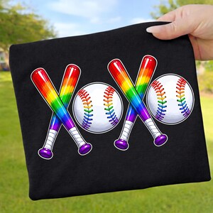 Könnte beinhalten: Ein schwarzes T-Shirt mit einem Grafikdesign aus zwei gekreuzten Baseballschläger-Paaren und zwei Baseballbällen, die das Wort XOXO bilden. Die Schläger und die Nähte der Bälle sind in einem regenbogenfarbenen Verlauf auf dem Stoff gestaltet.
