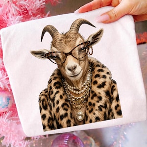 Könnte beinhalten: Ein weißes T-Shirt mit einer grafischen Illustration einer braunen Ziege, die eine Brille mit Leopardenmuster, einen passenden Kunstpelzmantel mit Leopardenmuster, eine mehrreihige Perlenkette und eine goldene Kette mit Kristallanhänger trägt.