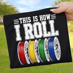 May include: Black t-shirt with the text "THIS IS HOW I ROLL" in white. Below the text are five spools of filament in red, yellow, green, and blue. The shirt is held up by a hand against a green background.